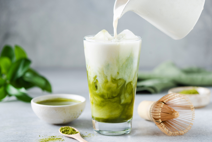 Milch wird in einen Iced Matcha Latte gegossen, daneben Matchapulver und ein Bambusbesen