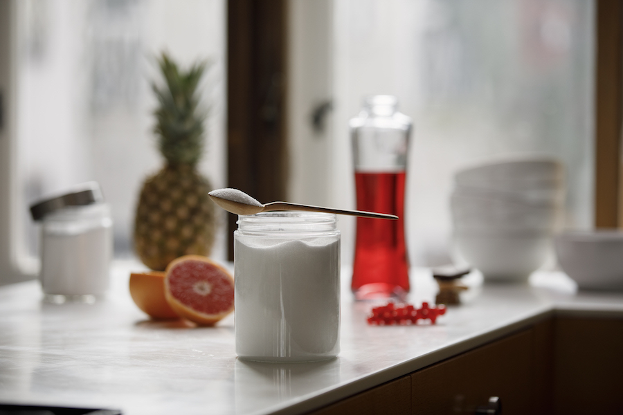 Glas mit weißem Zucker und Löffel auf einer Küchenzeile, Ananas, Grapefruit und roter Sirup im Hintergrund