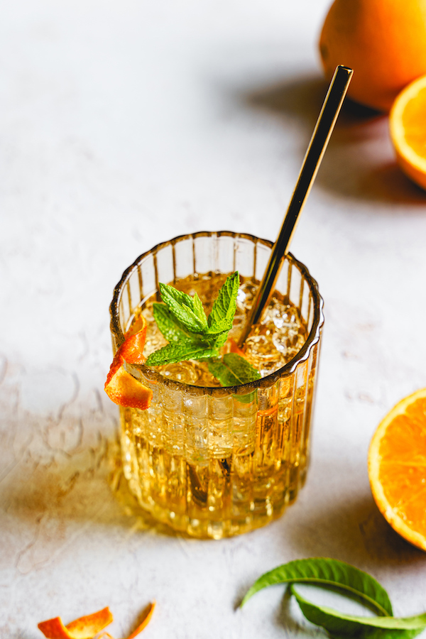 Eisgekühltes Orangengetränk im Rillenglas mit Minze, Orangenschale und Metallstrohhalm, Orangen daneben