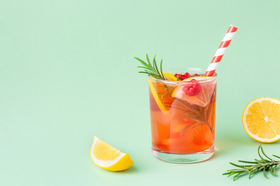 Glas roter Fruchtdrink mit Orange, Himbeeren, Rosmarin und gestreiftem Strohhalm auf mintgrünem Hintergrund