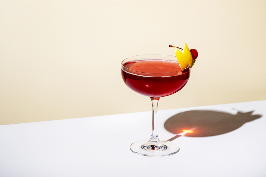 Roter Cocktail in einer Cocktailschale mit Zitronenzeste und Kirsche, Schatten auf weißem Tisch