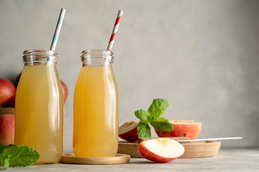 Zwei Flaschen naturtrüber Apfelsaft mit Papierstrohhalmen, Apfelspalten und Minze auf einem Tisch