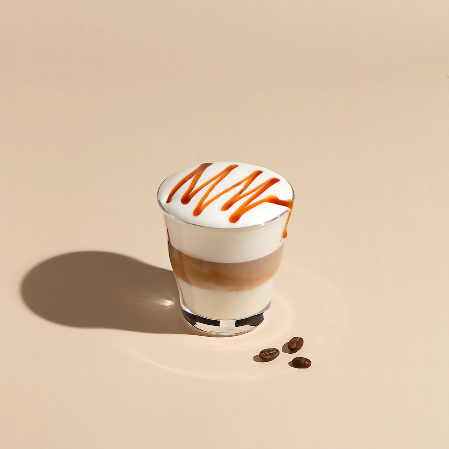 Latte Macchiato mit Karamellsauce im Glas neben Kaffeebohnen auf beigem Hintergrund