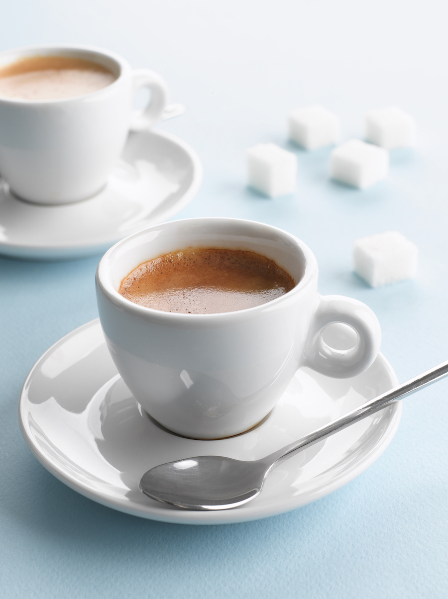 Zwei Tassen Espresso mit Zuckerwürfeln und einem Löffel auf hellblauem Tisch