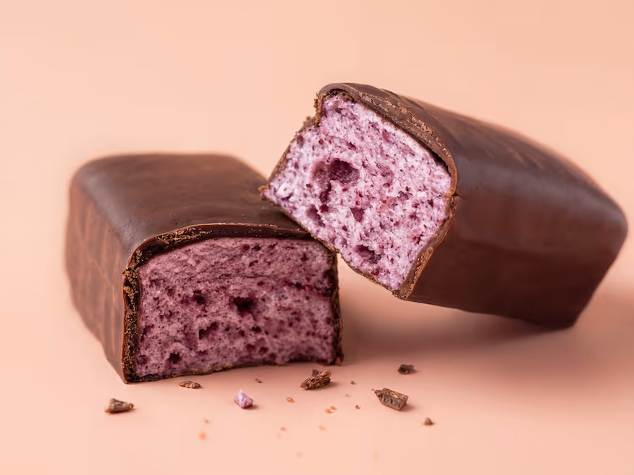 Zwei schokoladeüberzogene Riegel mit rosa Beeren-Schaumfüllung, einer aufgeschnitten, auf pfirsichfarbenem Hintergrund