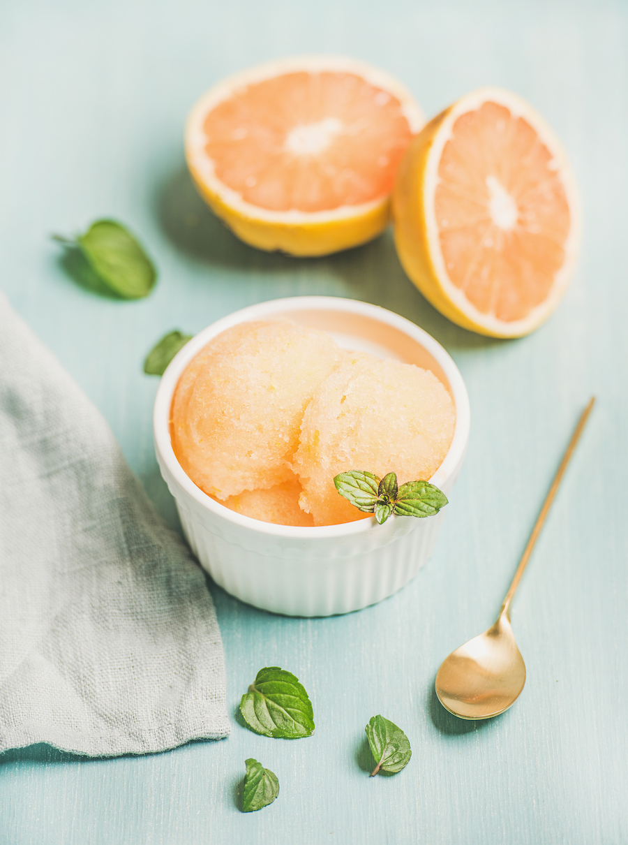 Grapefruit-Sorbet mit Minze in einer weißen Schale, halbierte Grapefruits und ein goldener Löffel auf hellblauem Tisch