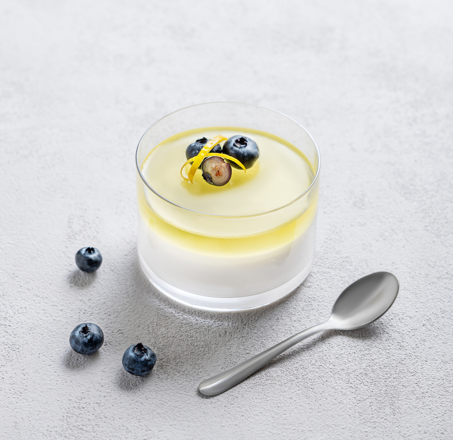 Glas Panna Cotta mit Zitronengelee, Heidelbeeren und Zitronenzeste, daneben ein Löffel