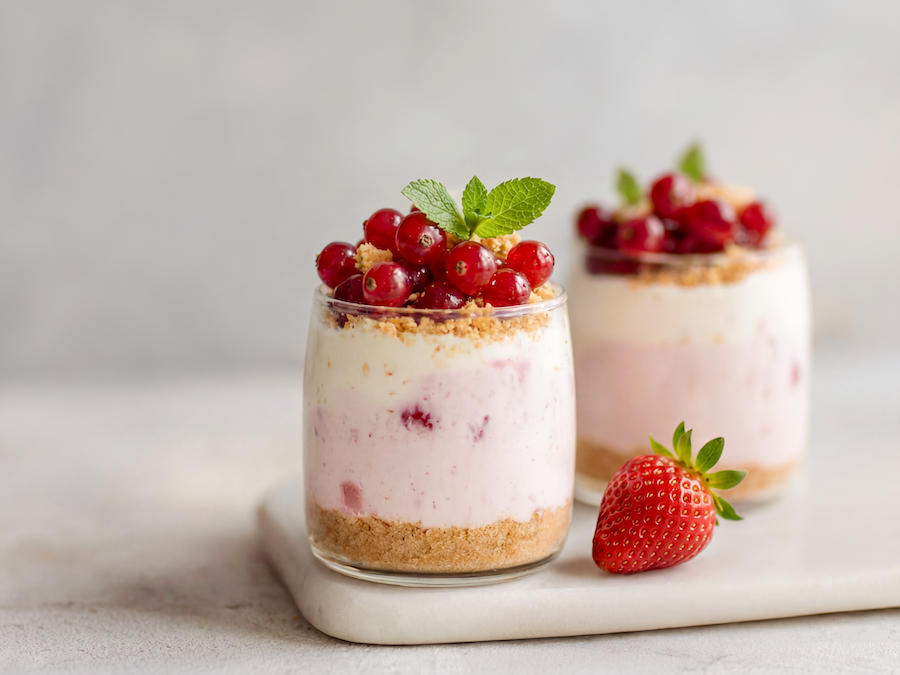 Zwei Gläser geschichtetes Erdbeer-Joghurt-Dessert mit Ribiseln und Minze, dazu eine frische Erdbeere