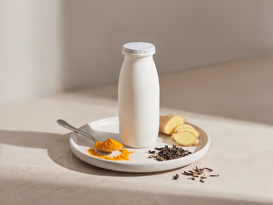 Weiße Trinkjoghurt-Flasche auf einem Teller mit Kurkumapulver, Ingwerscheiben und getrockneten Teeblättern