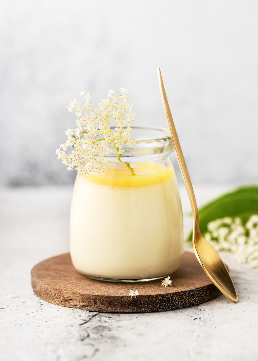 Glas Vanillepudding mit Holunderblüten und einem goldenen Löffel auf einem Holzbrett