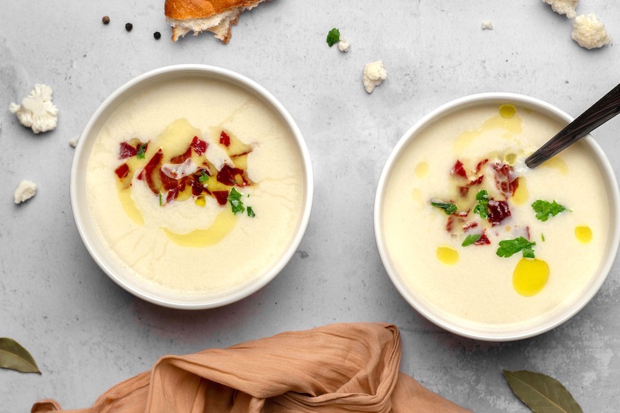 Zwei Schalen cremige weiße Suppe mit knusprigem Speck, Kräutern und Olivenöl, von oben