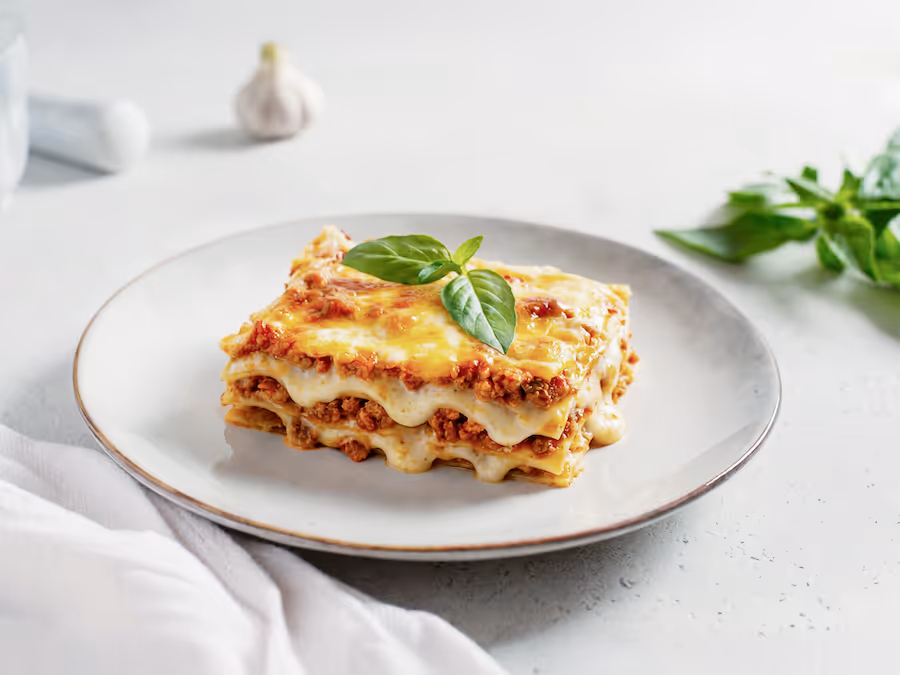 Stück Lasagne mit Faschiertem und geschmolzenem Käse auf einem Teller, garniert mit frischem Basilikum