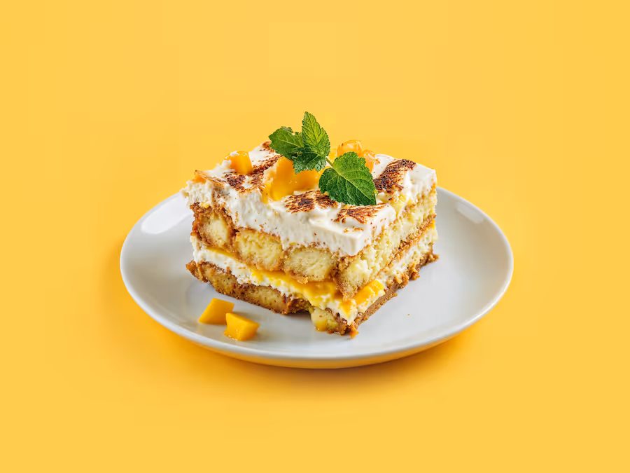 Stück Mango-Tiramisu mit Creme und Minzblättern auf einem Teller vor gelbem Hintergrund
