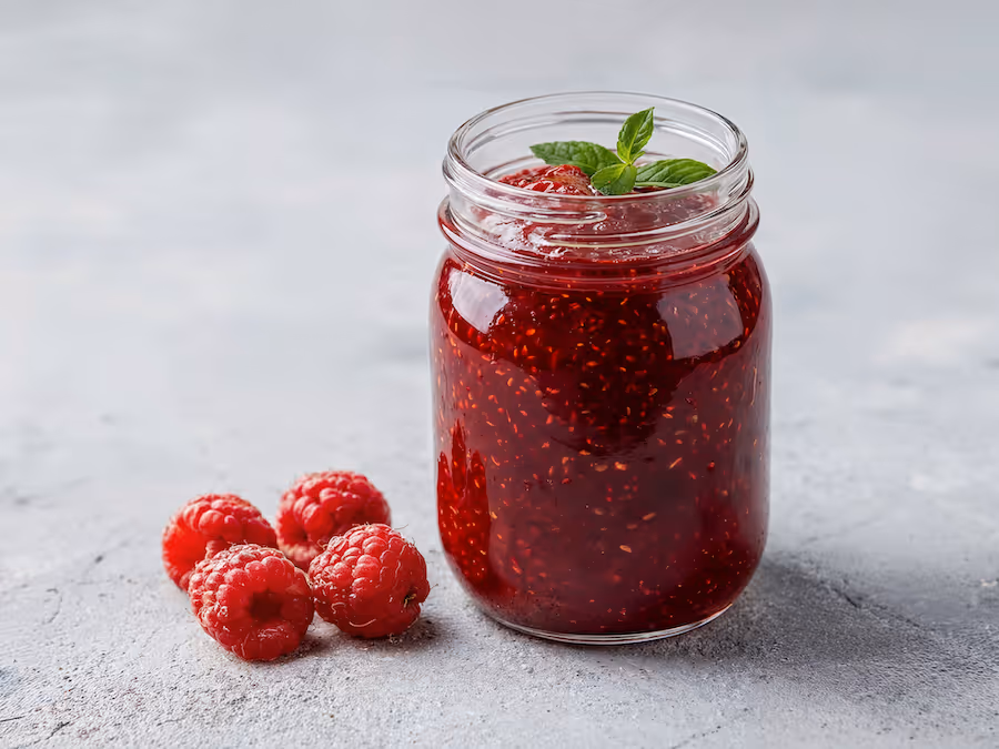 Glas Himbeermarmelade mit Minzblatt und frischen Himbeeren auf grauer Fläche