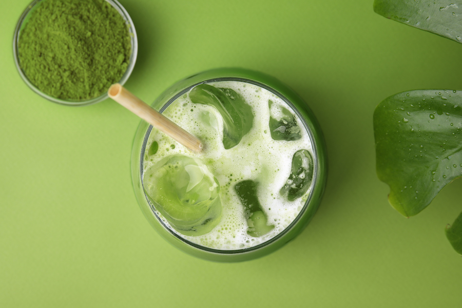 Iced Matcha Latte mit Trinkhalm neben einer Schale Matchapulver vor grünem Hintergrund