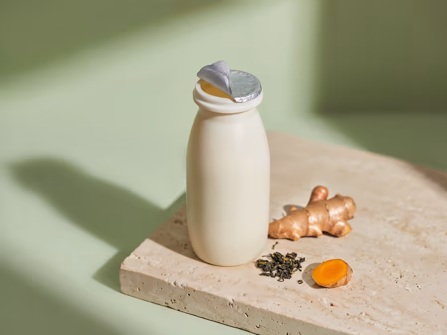 Trinkjoghurt-Flasche mit Foliendeckel auf einem Steinbrett mit frischem Ingwer, Kurkuma und Teeblättern