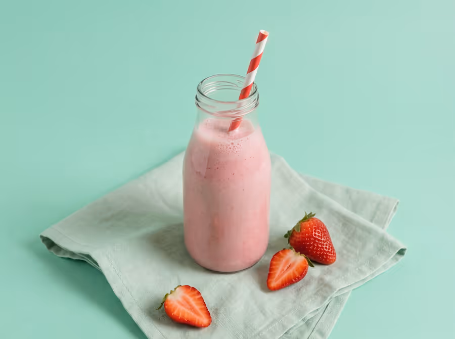 Flasche Erdbeer-Smoothie mit gestreiftem Trinkhalm und halbierten Erdbeeren auf mintgrünem Tuch