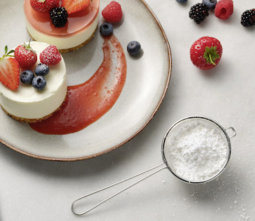 Mini-Cheesecakes mit Beeren und Fruchtsauce auf einem Teller, dazu ein Sieb mit Staubzucker