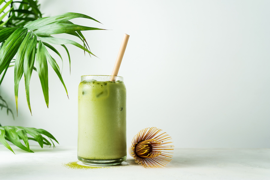 Iced matcha latte in a glass with a straw, next to a bamboo whisk and palm leaves