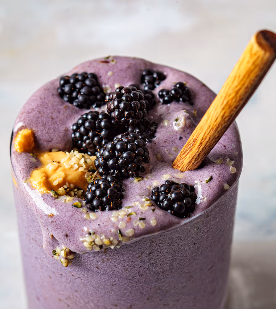 Violetter Brombeer-Smoothie mit frischen Brombeeren, Nussmus und Hanfsamen, dazu ein Holzlöffel