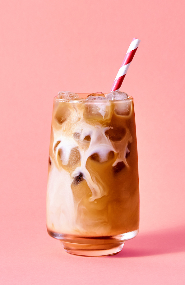 Glas Eiskaffee mit Milchschlieren und rot gestreiftem Trinkhalm vor rosa Hintergrund