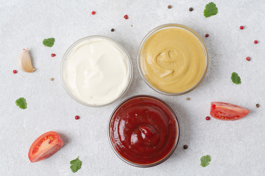 Drei Schälchen mit Mayonnaise, Senf und Ketchup, dazu Tomatenspalten, Knoblauch und Kräuter von oben