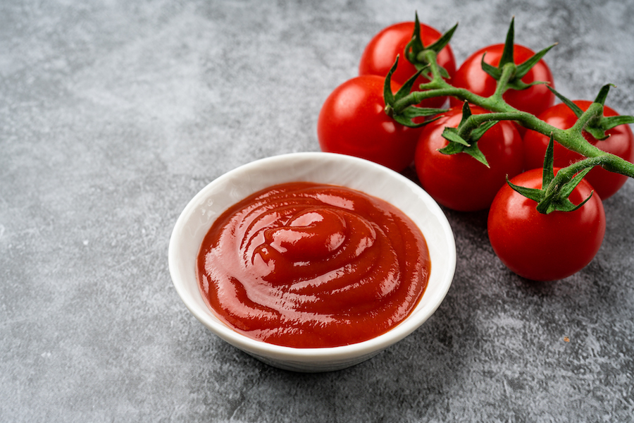 Weiße Schale mit Ketchup neben Rispentomaten auf grauem Stein