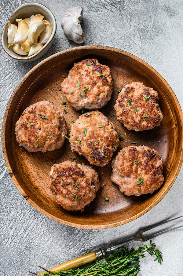 Sechs gebratene Fleischlaibchen mit Kräutern in einer Holzschale, daneben Knoblauchzehen und Thymian