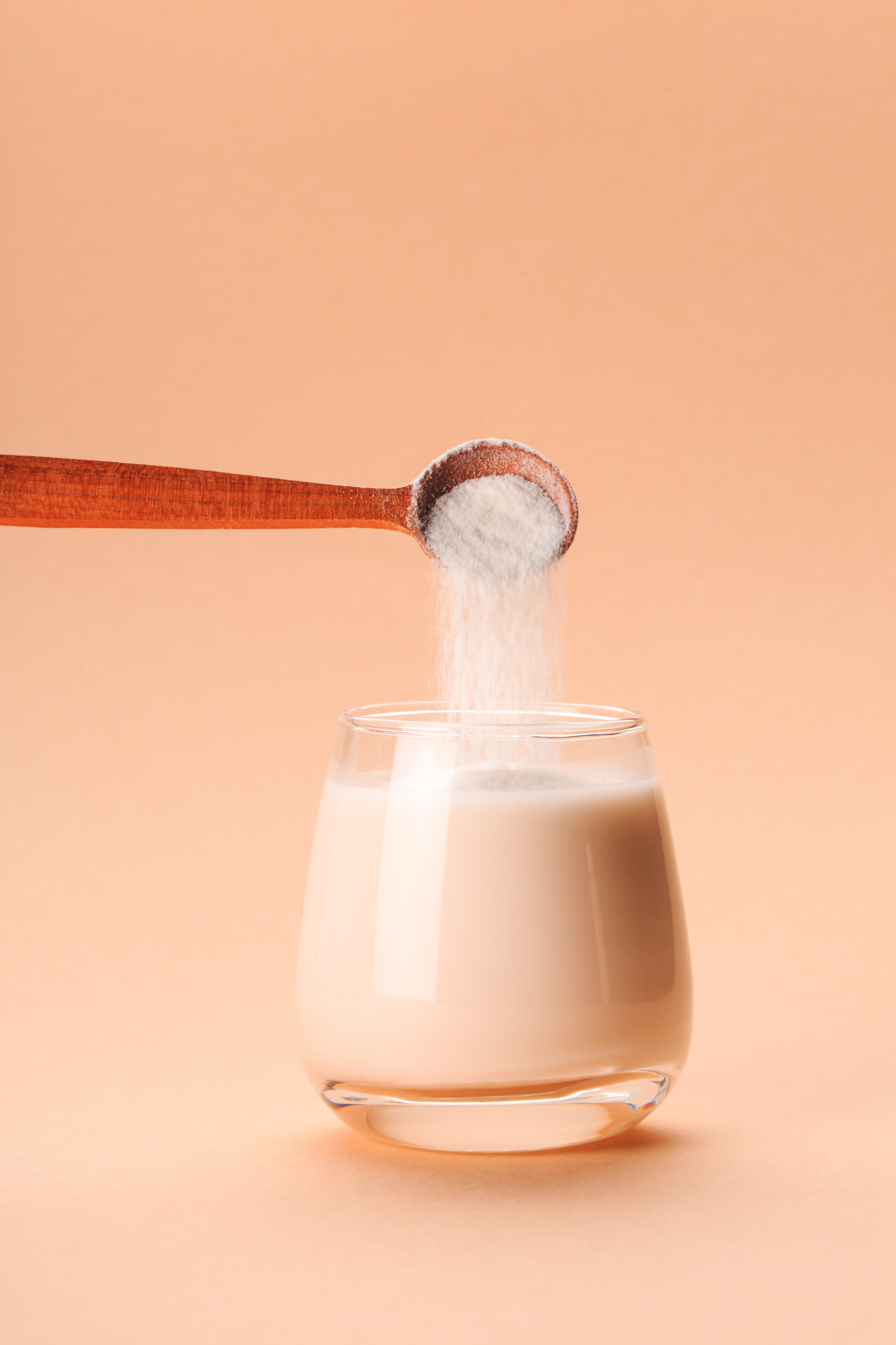 Pulver rieselt von einem Holzlöffel in ein Glas Milch vor pfirsichfarbenem Hintergrund
