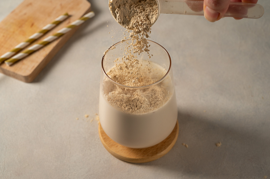 Proteinpulver wird aus einem Messlöffel in ein Glas Milch auf einem Holzuntersetzer geschüttet