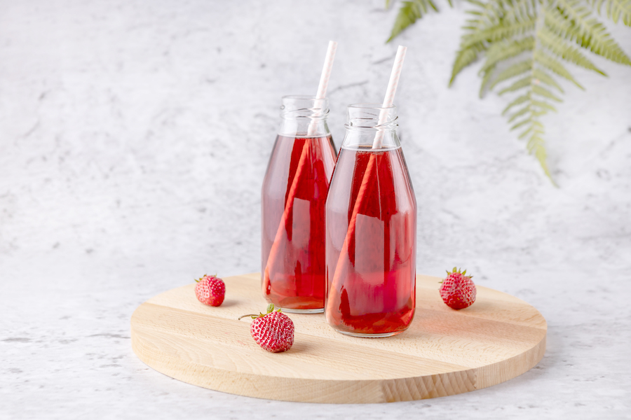 Zwei Glasflaschen roter Erdbeersaft mit Strohhalmen und frischen Erdbeeren auf einem runden Holzbrett