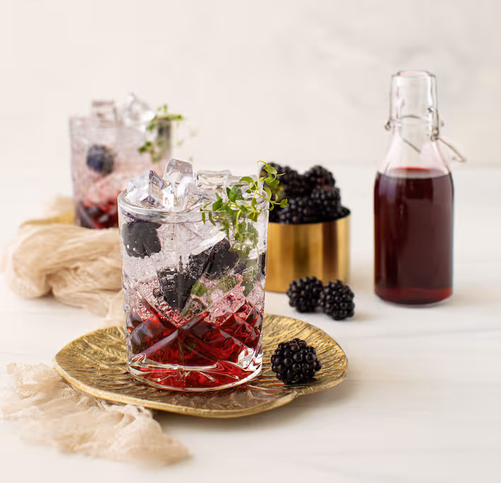 Zwei Gläser Brombeer-Spritz mit Eis und Thymian neben einer Flasche dunklem Beerensirup