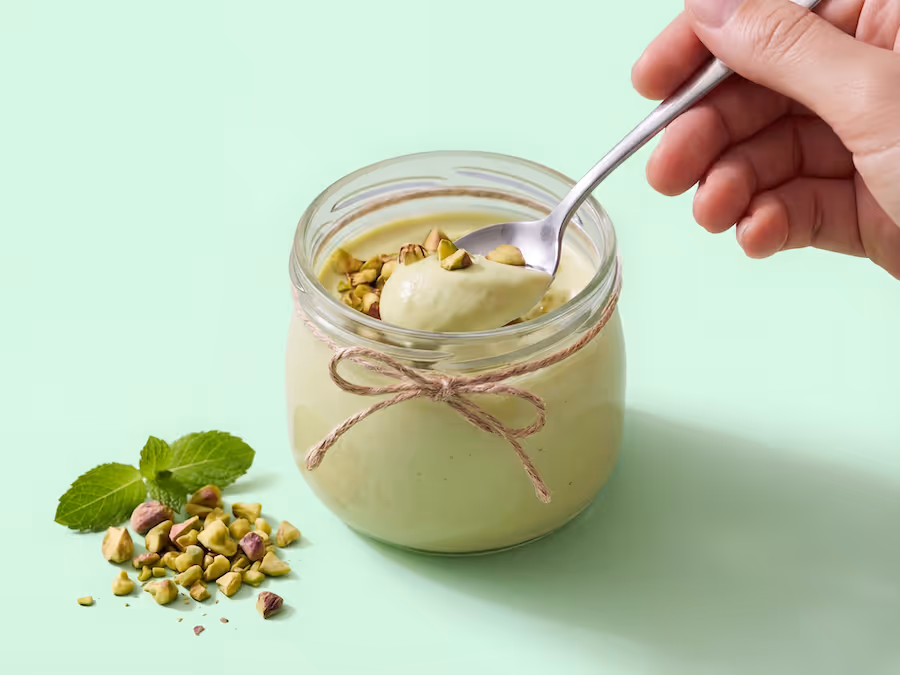 Glas Pistaziencreme mit Löffel, gehackten Pistazien und Minzblättern auf hellgrünem Hintergrund