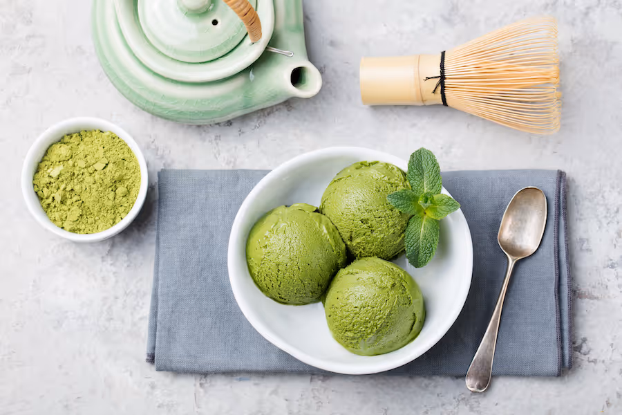 Drei Kugeln Matcha-Eis in einer Schale mit Minze, daneben Matchapulver, Besen und Teekanne