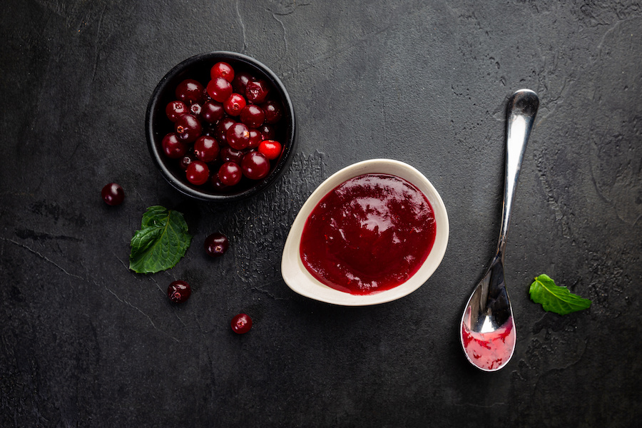 Schälchen mit roter Beerenkonfitüre, Cranberrys, Minzblättern und Löffel auf dunklem Schiefer von oben