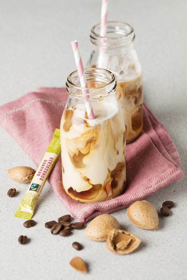 Zwei Flaschen Eiskaffee mit gestreiften Strohhalmen, ein Stick brauner Rohrzucker, Kaffeebohnen und Mandeln