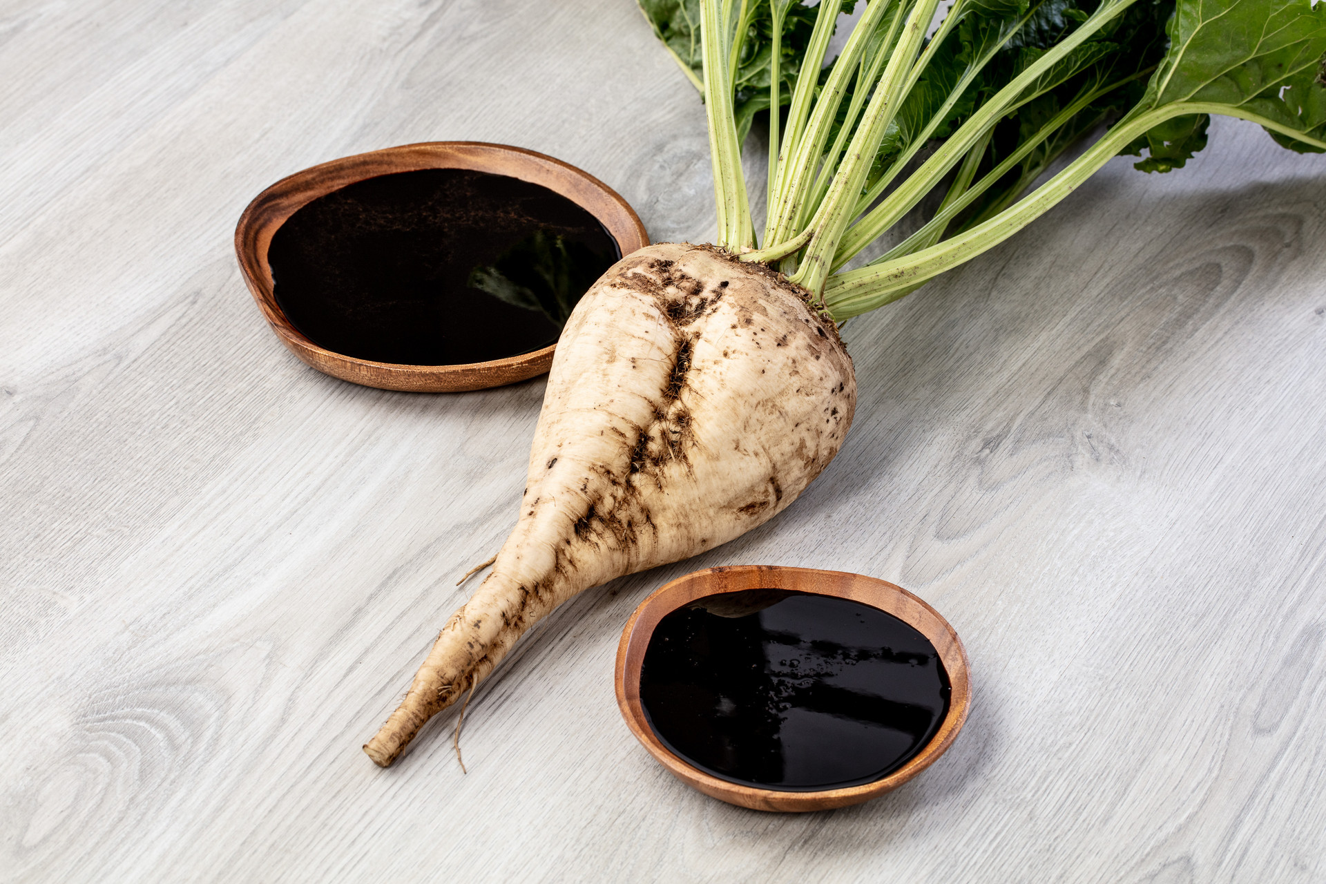 Zuckerrübe mit grünen Blättern auf einem Holztisch neben zwei Holzschalen mit dunklem Rübensirup