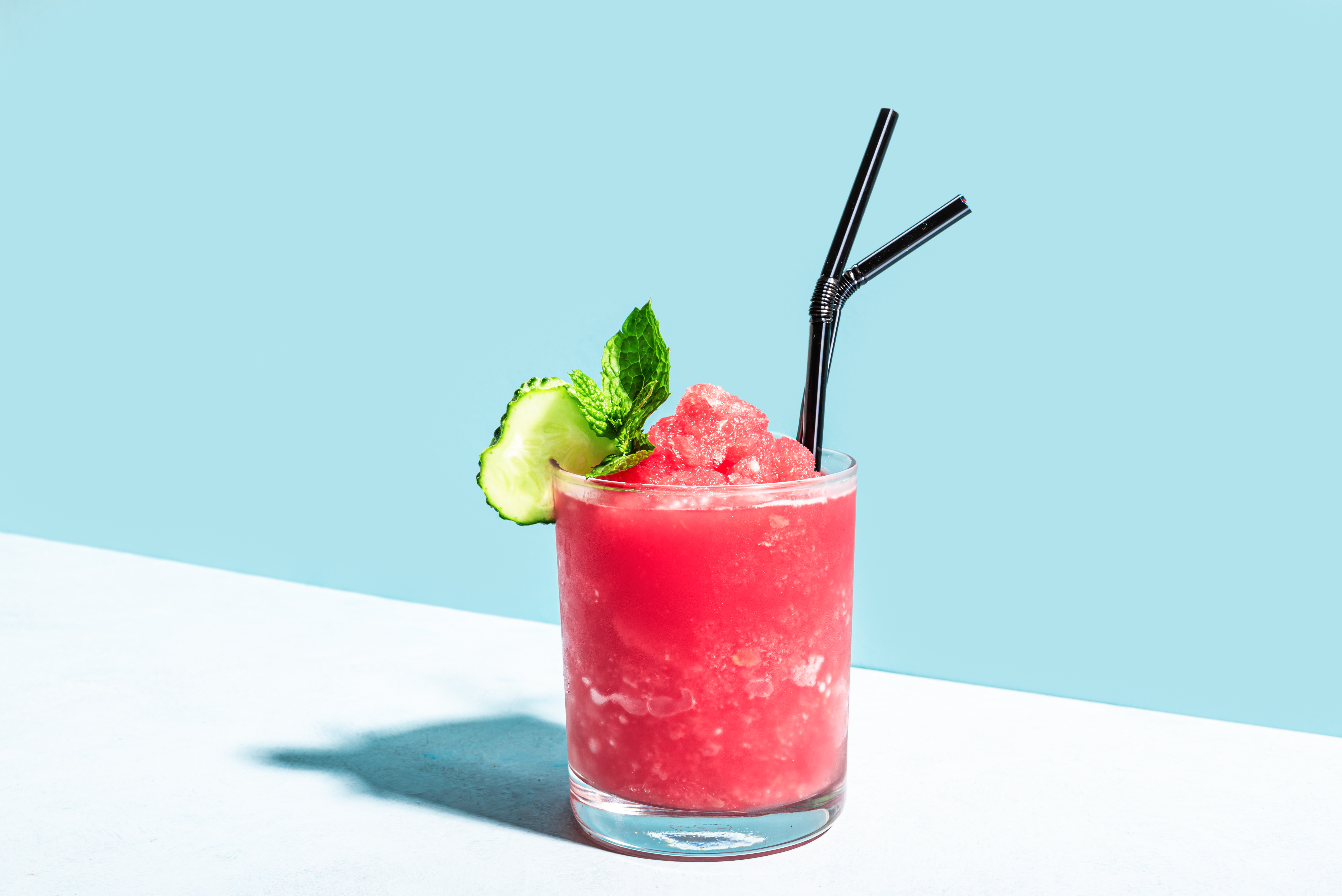 Wassermelonen-Slush im Glas mit Gurke, Minze und schwarzen Strohhalmen vor hellblauem Hintergrund