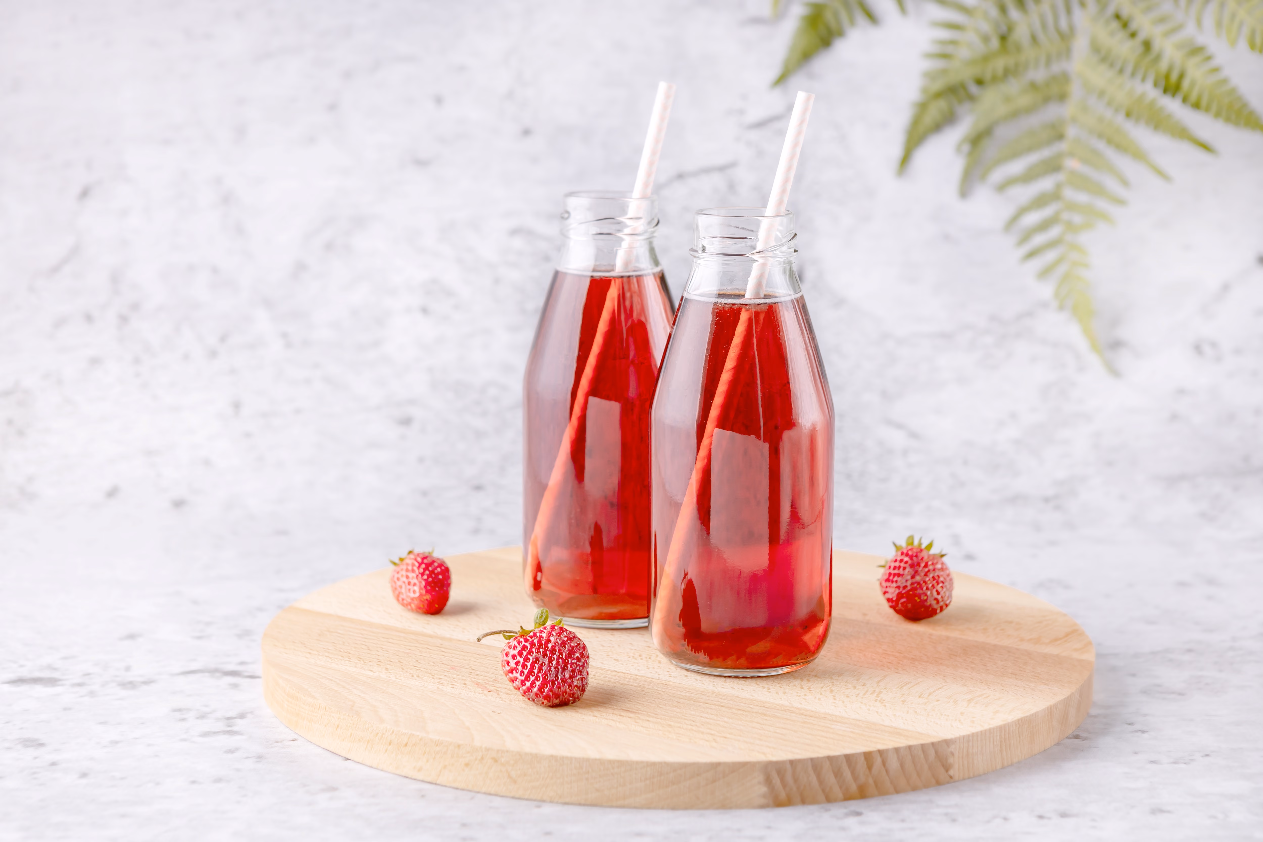 Zwei Glasflaschen roter Erdbeersaft mit Strohhalmen und frischen Erdbeeren auf einem runden Holzbrett