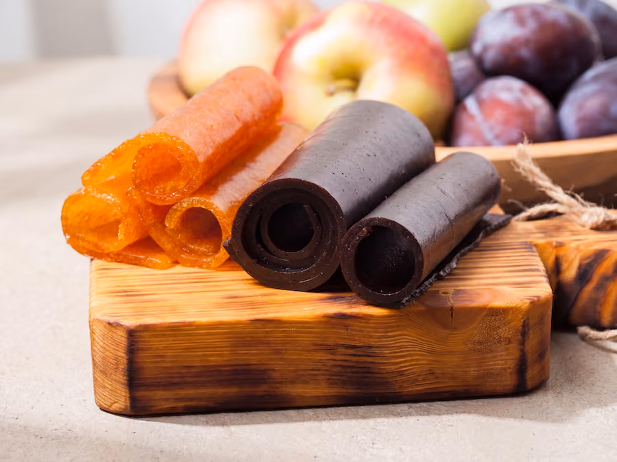 Rolled apricot and plum fruit leather on a wooden board with fresh apples and plums behind