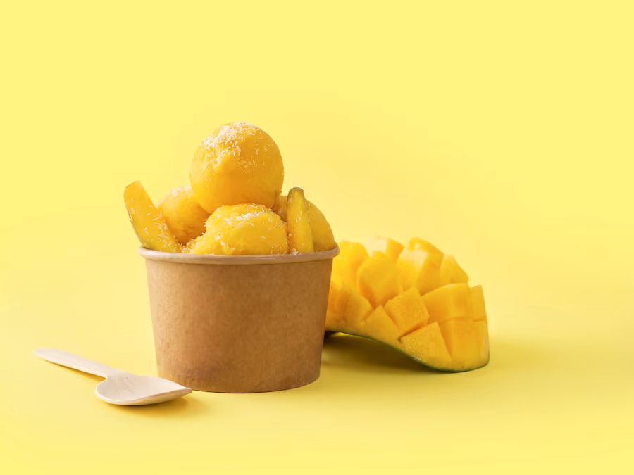 Paper cup of frozen mango puree scoops next to a diced mango on a yellow background