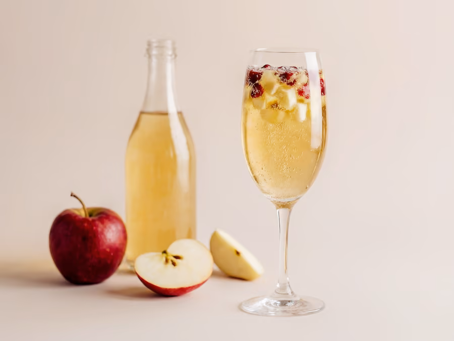 Glas prickelnder Cider mit Beeren neben einer Flasche und frischen Äpfeln