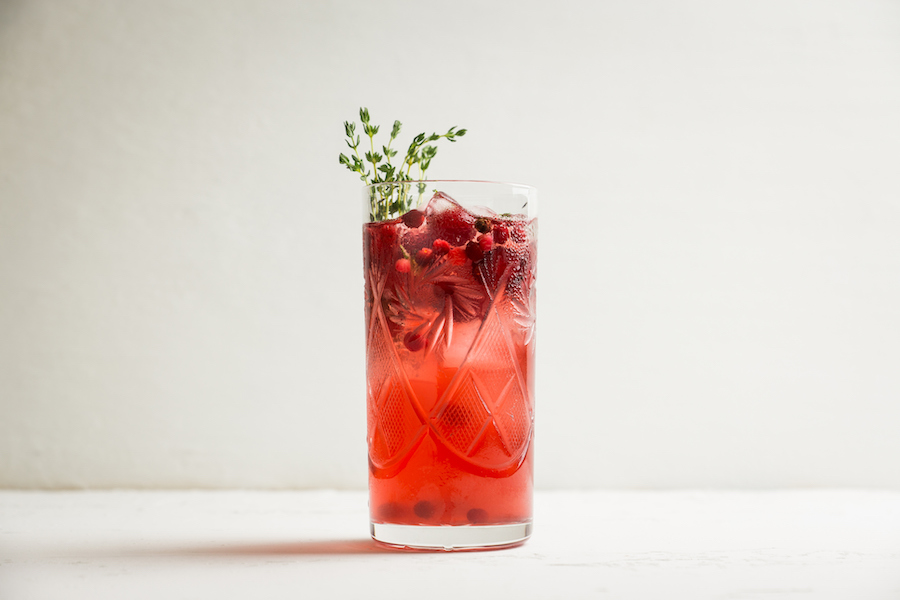 Hohes Glas mit rotem Beerengetränk, Eis, Cranberrys und einem Thymianzweig auf weißem Tisch