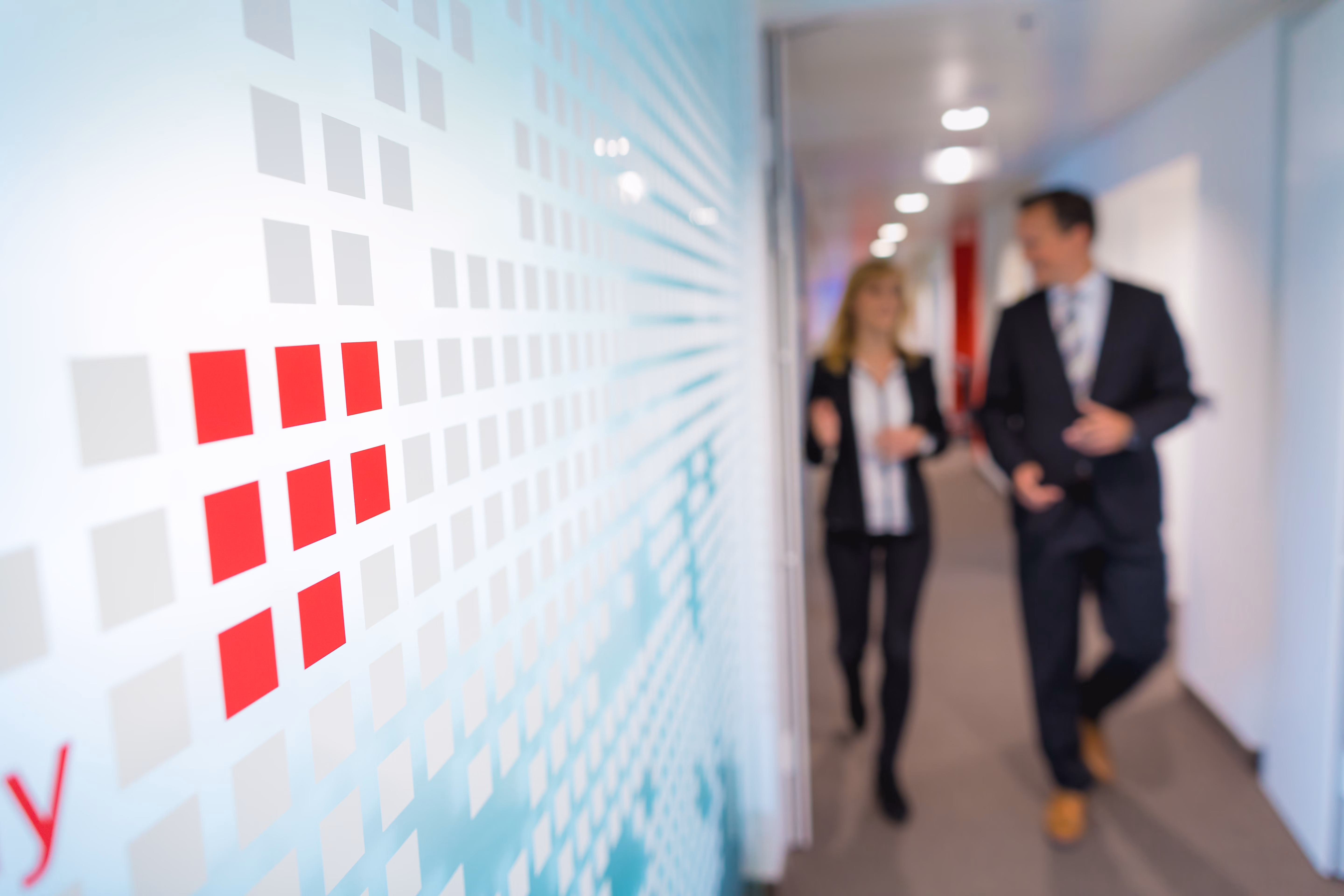 Zwei Mitarbeitende gehen durch einen Bürogang an einer Wand mit rot-grauem Quadratmuster vorbei