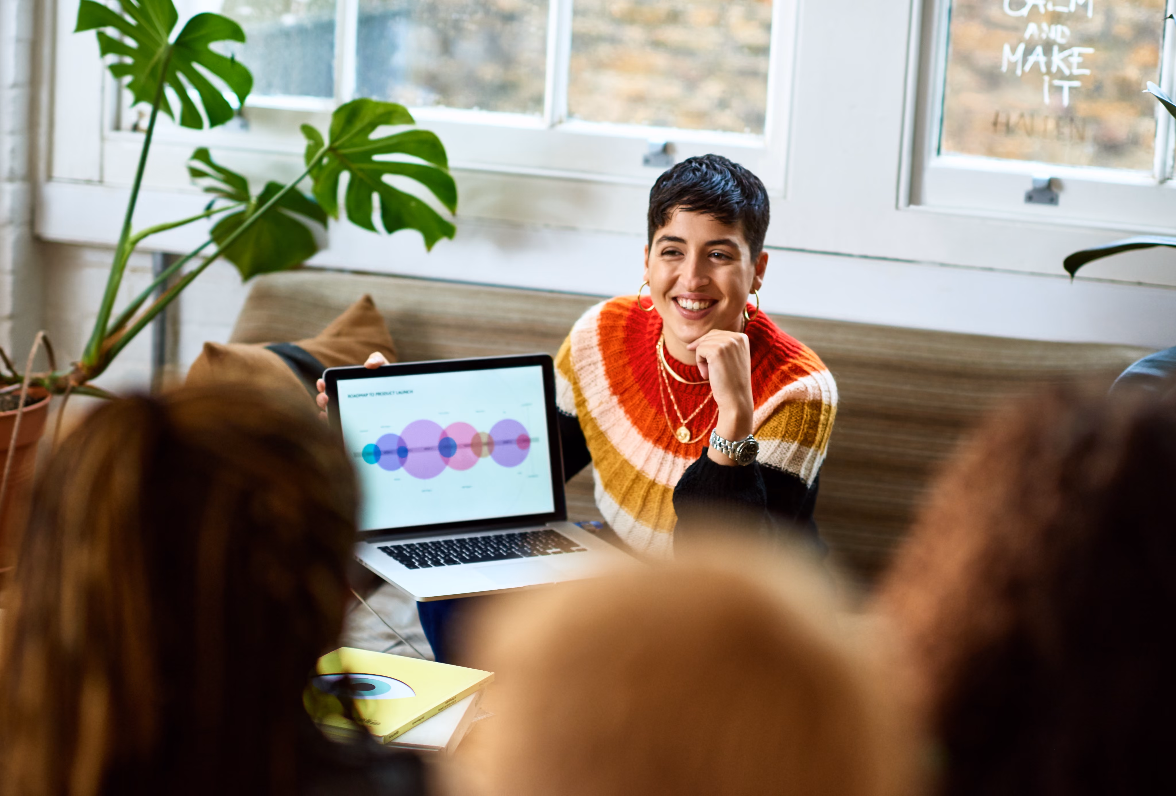 Smiling young woman presenting a chart on a laptop to a small group in a bright office [no AI]