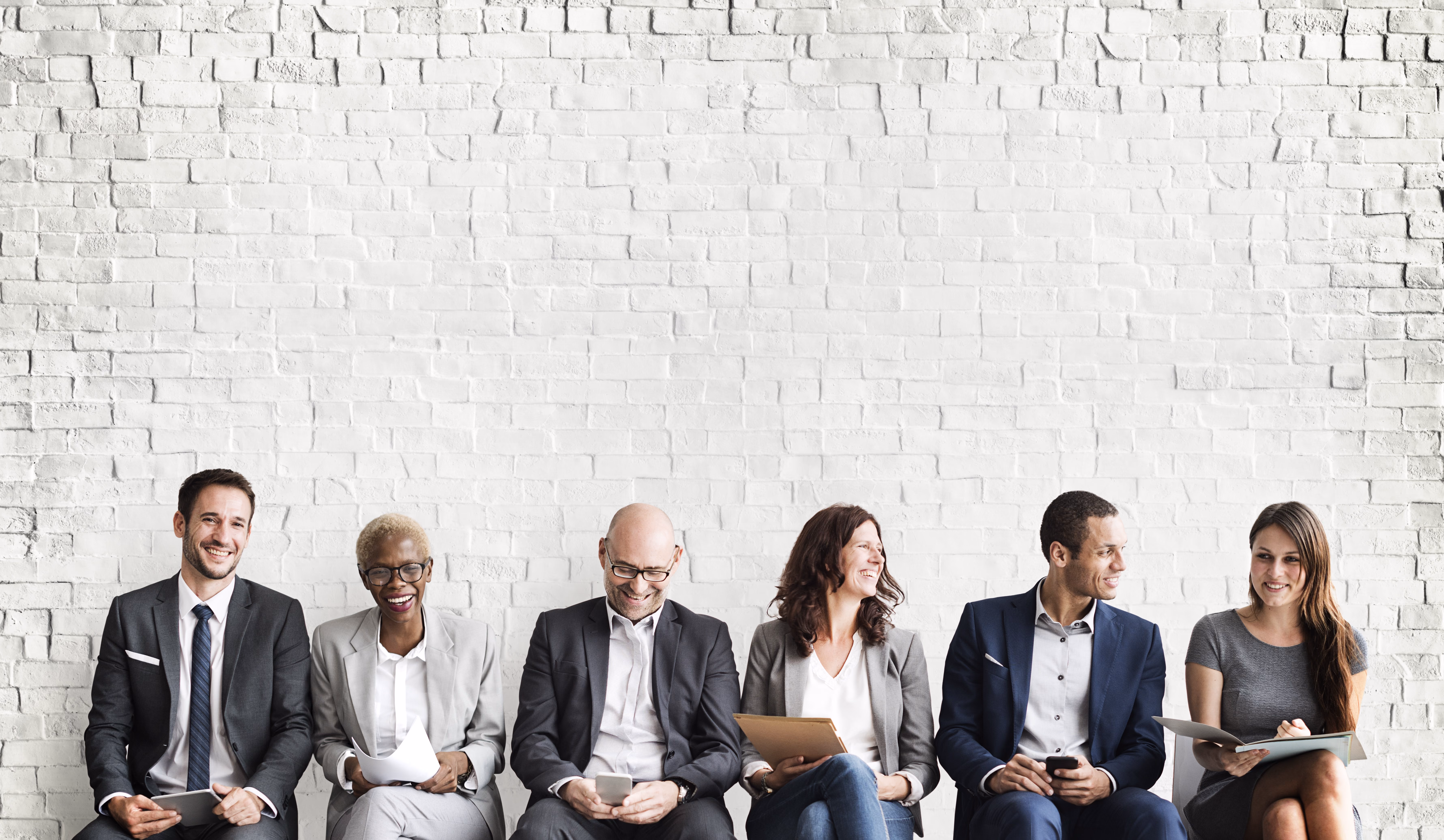 Six smiling applicants in business attire sitting in a row in front of a white brick wall