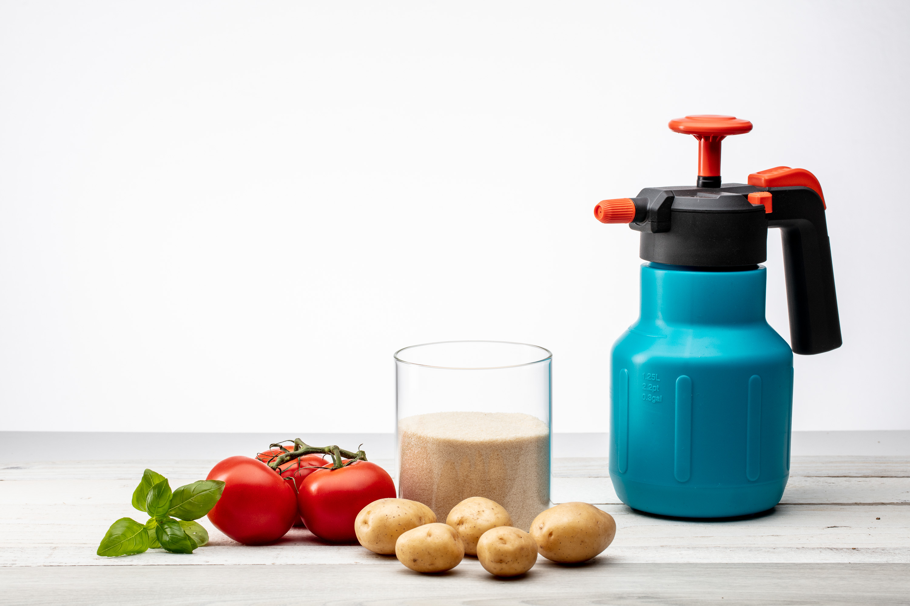 Drucksprüher und ein Glas Düngergranulat neben Tomaten, Kartoffeln und Basilikum