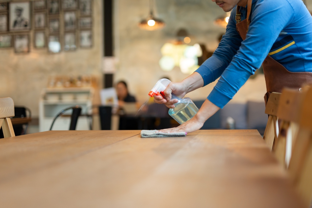 Person in an apron wiping a wooden café table with a cloth and spray bottle