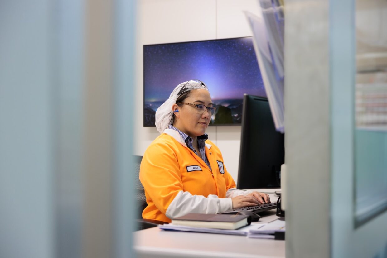 Employee in an orange work jacket and hairnet working at a computer in the plant office [no AI]