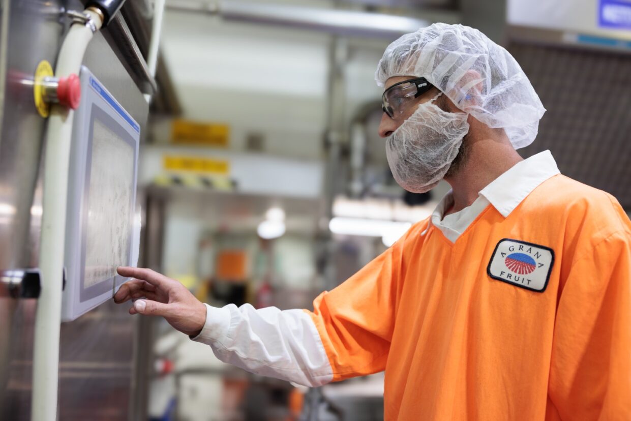 Employee in an orange AGRANA Fruit jacket and hairnet operating a touchscreen control panel